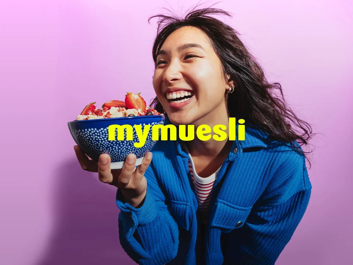 Eine lachende Frau in einer blauen Jacke hält eine blaue Schale Müsli mit Erdbeeren vor einem lila Hintergrund. Der gelbe Text 'mymuesli' ist überlagert.