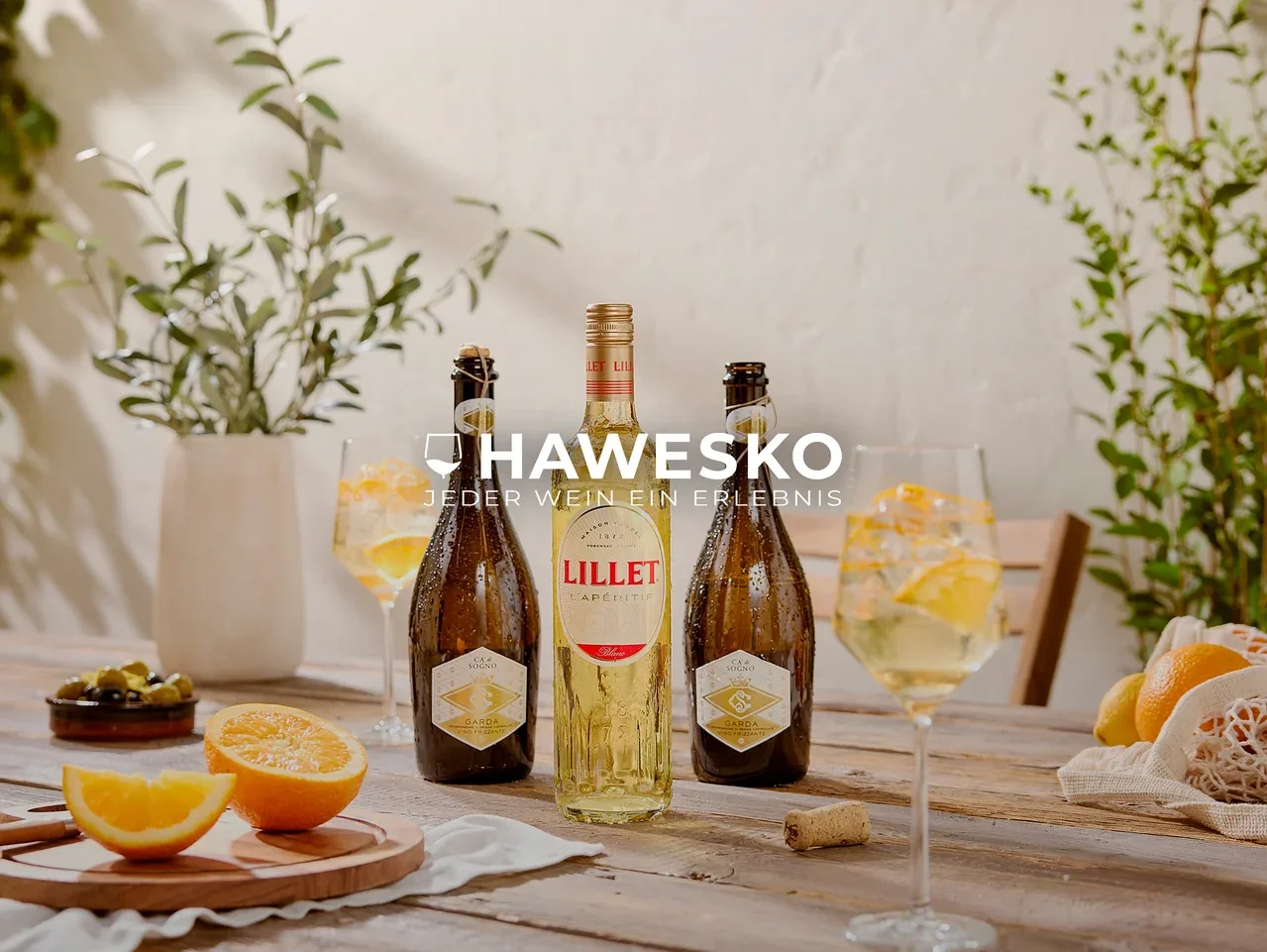 Drei kalte Flaschen, eine Lillet und zwei Garda-Schaumweine, stehen auf einem rustikalen Holztisch. Daneben sind zwei Gläser mit Aperitifs, Eis und Orangenscheiben, sowie geschnittene Orangen, grüne Oliven und ganze Zitrusfrüchte. Grüne Pflanzen befinden sich im Hintergrund.