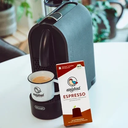 Eine Tasse Kaffee steht auf einer DeLonghi Maschine, daneben eine Packung earlybird Bio Espresso Kapseln.