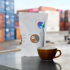 Eine weiße Packung Hermetic Coffee Roasters Kaffee steht neben einer braunen Tasse vor dem Hintergrund von Containern.