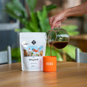 Eine Hand gießt Kaffee aus einer Kanne in eine orangefarbene Tasse neben einer Packung Alltagsheld-Kaffee.