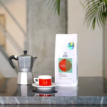 Auf einem Tisch stehen eine Kaffeemaschine, eine Tasse und eine Packung Black Delight x roasted. - Momento Italiano Co-Roast #2 Espresso.