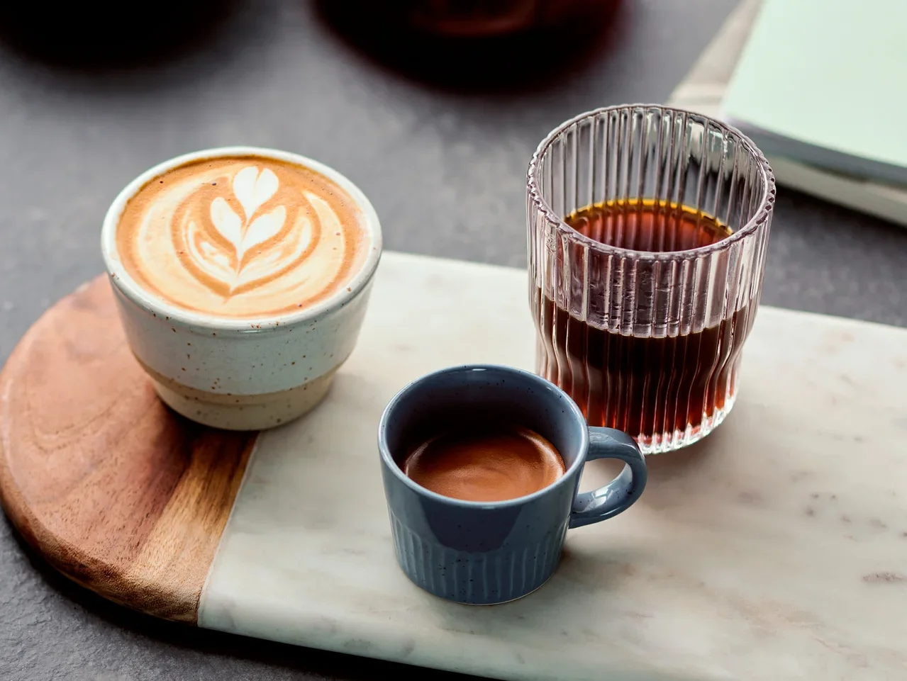 Auf einem Marmorbrett stehen ein Cappuccino, ein Espresso in einer blauen Tasse und ein Glas Kaffee.