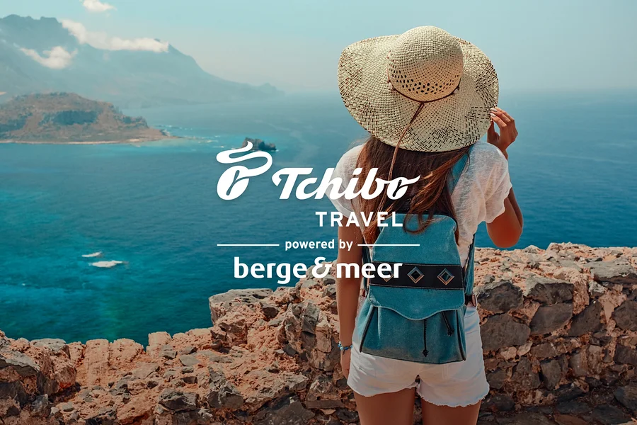 Frau mit Hut und blauem Rucksack steht auf Felsen mit Blick aufs Meer. Logos von Tchibo Travel und Berge&Meer.
