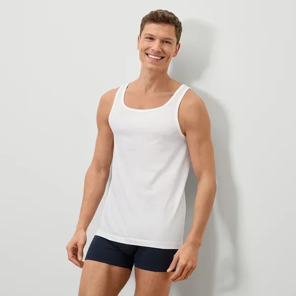 Ein lächelnder Mann mit kurzem braunem Haar trägt ein weißes Tanktop und blaue Boxershorts vor einer weißen Wand.