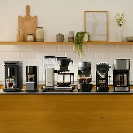Sechs verschiedene Kaffeemaschinen stehen auf einer dunklen Kücheninsel-Arbeitsplatte. Von links nach rechts sind zwei schwarze Einzelportionsmaschinen, eine silber-schwarze Filterkaffeemaschine, eine dunkelgraue Maschine mit zwei Tassen, eine schwarze Espressomaschine und eine weitere silber-schwarze Filterkaffeemaschine zu sehen. Einige Maschinen haben Tassen mit Kaffee darunter. Im Hintergrund befindet sich ein Holzregal mit Küchendekoration.