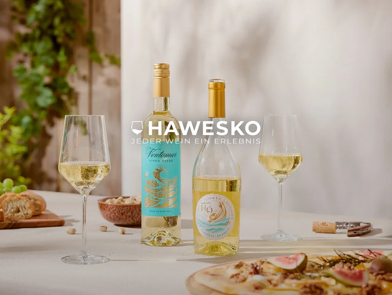 Zwei Flaschen Weißwein und zwei Gläser Weißwein auf einem Tisch mit Fladenbrot, Pistazien, Brot und Trauben. Im Hintergrund ist grünes Laub zu sehen. Text: HAWESKO JEDER WEIN EIN ERLEBNIS.