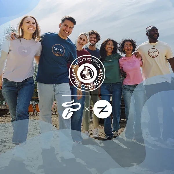 Sechs lächelnde Personen, drei Männer und drei Frauen, stehen Arm in Arm an einem Sandstrand. Sie tragen verschiedenfarbige HOOD.-T-Shirts und Jeans und blicken nach rechts. Ein „Wir unterstützen Viva con Agua“-Logo ist zentriert.