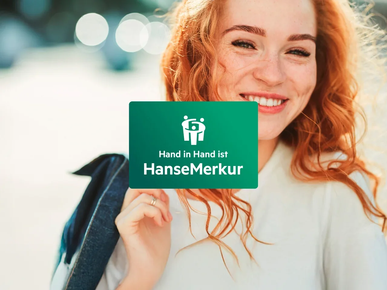 Eine lächelnde rothaarige Frau hält eine grüne Karte mit dem Logo und dem Schriftzug HanseMerkur.