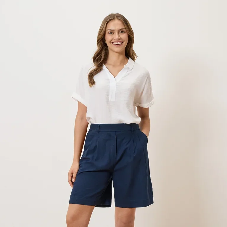 Frau mit weißer Bluse und blauen Shorts steht vor einem weißen Hintergrund.