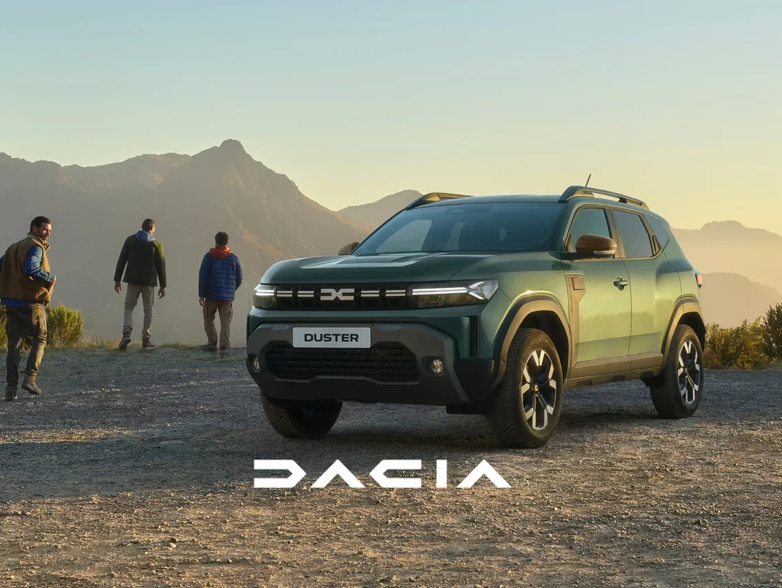 Ein grüner Dacia Duster SUV steht auf einem Schotterweg. Drei Männer stehen dahinter und blicken auf eine weite Berglandschaft im warmen Licht des Sonnenuntergangs. Unten ist das Dacia Logo zu sehen.