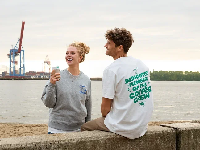 Frau lacht und hält einen Kaffeebecher in der Hand, Mann sitzt auf einer Mauer am Strand.