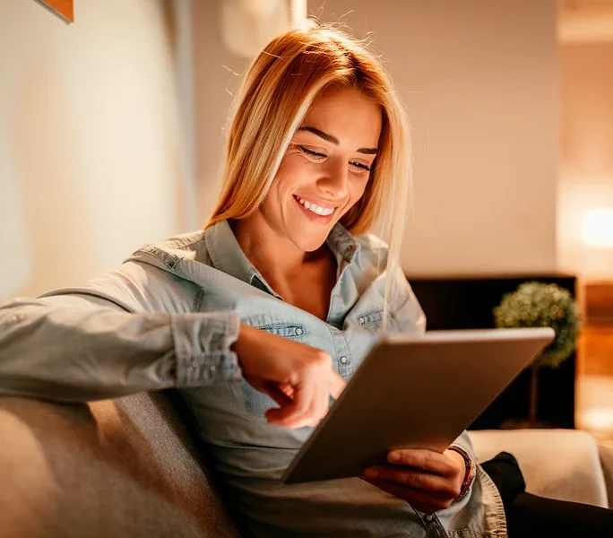 Frau lächelt und benutzt ein Tablet, während sie auf einem Sofa sitzt.