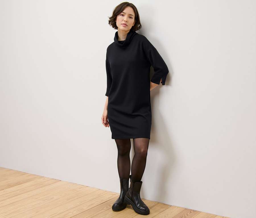 Frau steht an der Wand und trägt ein Sweatkleid aus Modal-Mix, eine schwarze Strumpfhose und schwarze Stiefel.