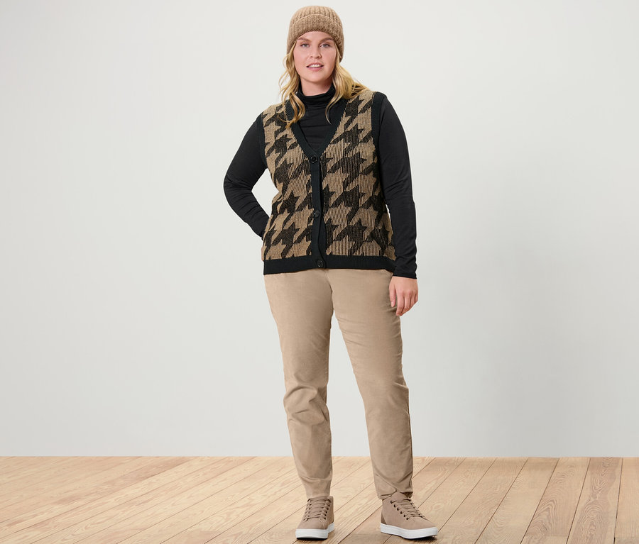 Eine Frau steht und trägt ein schwarzes Rollkragenshirt, eine Strickweste mit Hahnentritt-Jacquard und eine camel Samthose.