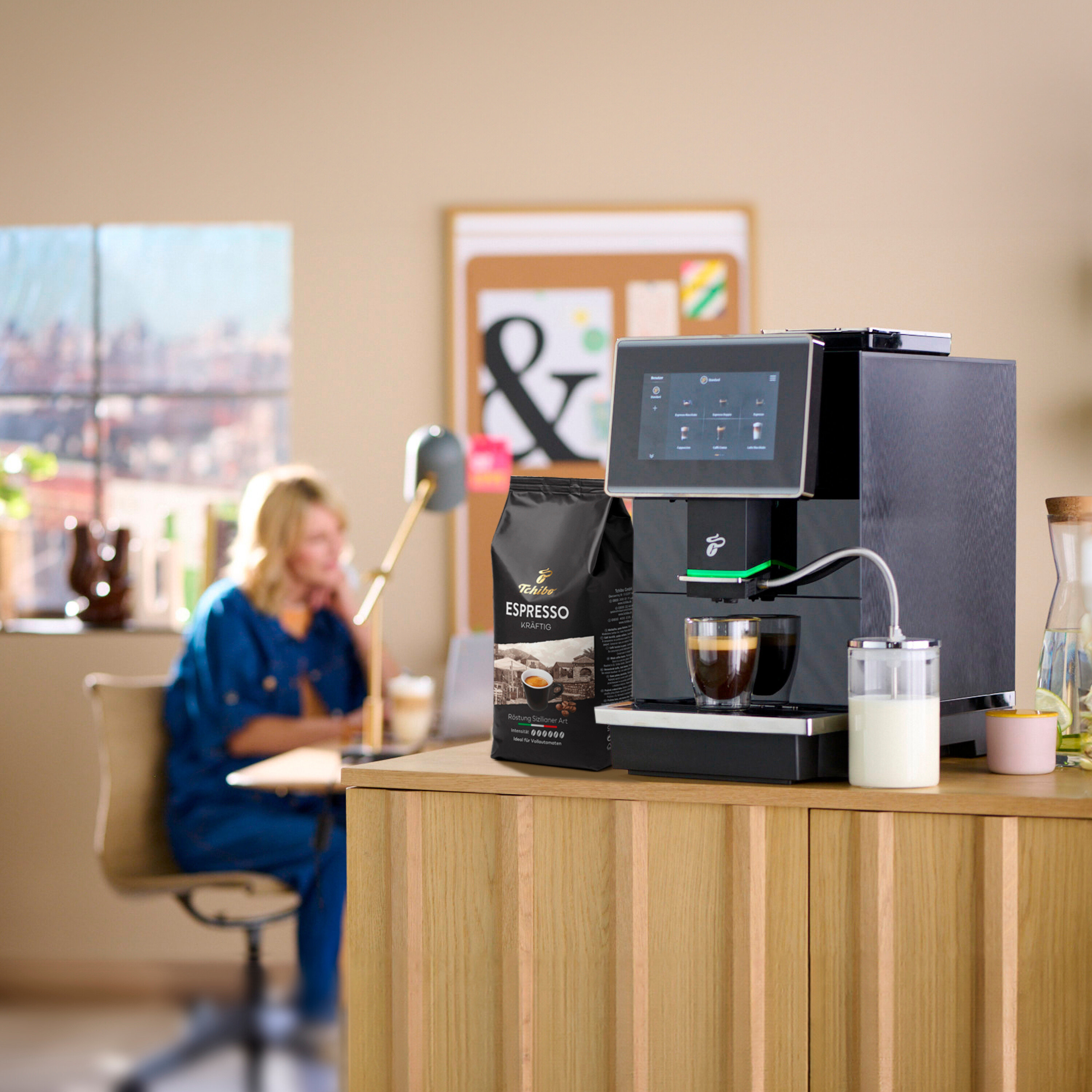 Eine Frau sitzt an einem Tisch im Hintergrund. Im Vordergrund steht eine Tchibo Espresso Maschine mit einer Tasse Kaffee und einer Packung Espresso.