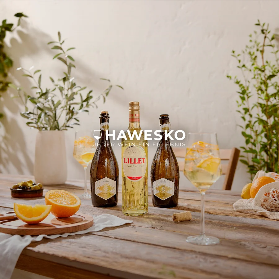 Drei kalte Flaschen, eine Lillet und zwei Garda-Schaumweine, stehen auf einem rustikalen Holztisch. Daneben sind zwei Gläser mit Aperitifs, Eis und Orangenscheiben, sowie geschnittene Orangen, grüne Oliven und ganze Zitrusfrüchte. Grüne Pflanzen befinden sich im Hintergrund.