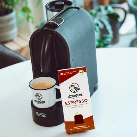 Eine Tasse Kaffee steht auf einer DeLonghi Maschine, daneben eine Packung earlybird Bio Espresso Kapseln.