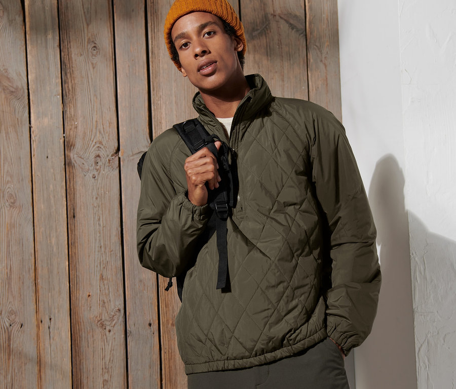 Mann posiert vor einer Holzwand, trägt eine Windprotection-Jacke und eine gelbe Mütze.