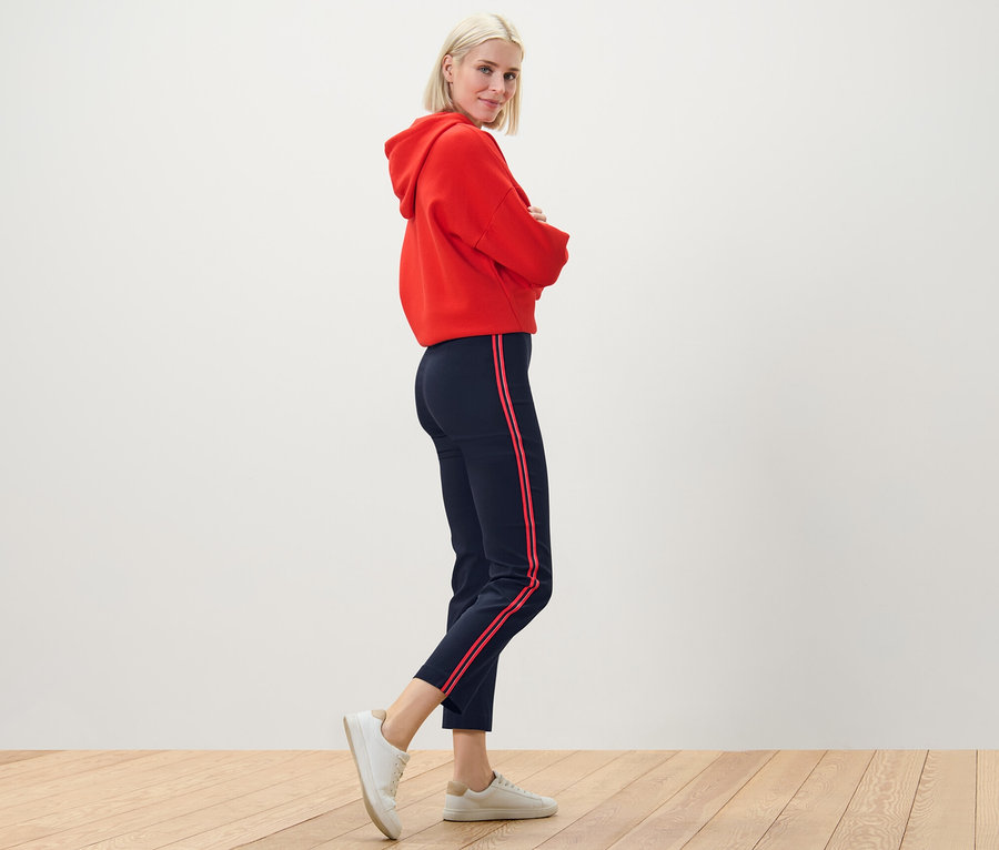 Blonde Frau posiert in rotem Hoodie und Stretchhose mit Galonstreifen.