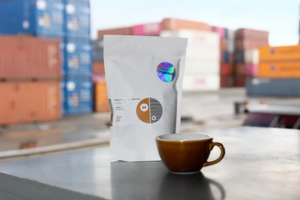Eine weiße Packung Hermetic Coffee Roasters Kaffee steht neben einer braunen Tasse vor dem Hintergrund von Containern.