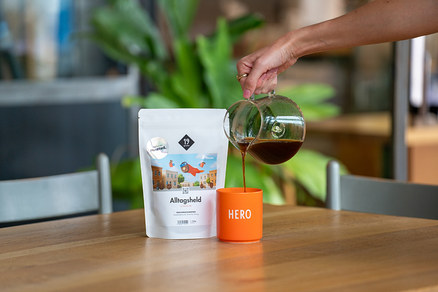 Eine Hand gießt Kaffee aus einer Kanne in eine orangefarbene Tasse neben einer Packung Alltagsheld-Kaffee.
