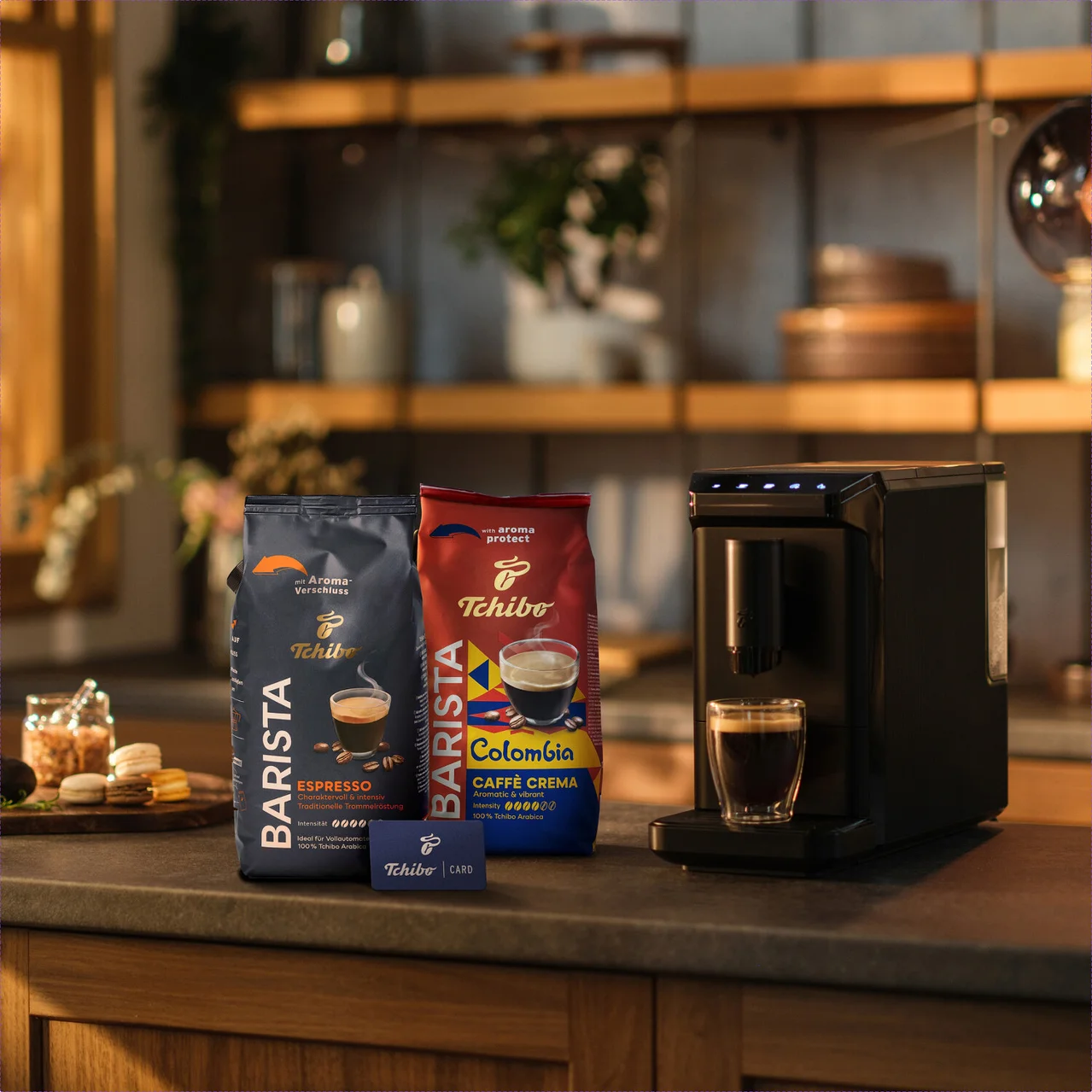 Zwei Packungen Tchibo Barista Kaffee, Espresso und Colombia Caffè Crema, stehen neben einer schwarzen Kaffeemaschine mit einer Tasse Kaffee und einer Tchibo Card auf einer Küchenzeile.