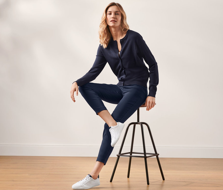Blonde Frau sitzt auf einem Hocker und trägt eine dunkelblaue Stretchhose und eine Strickjacke.