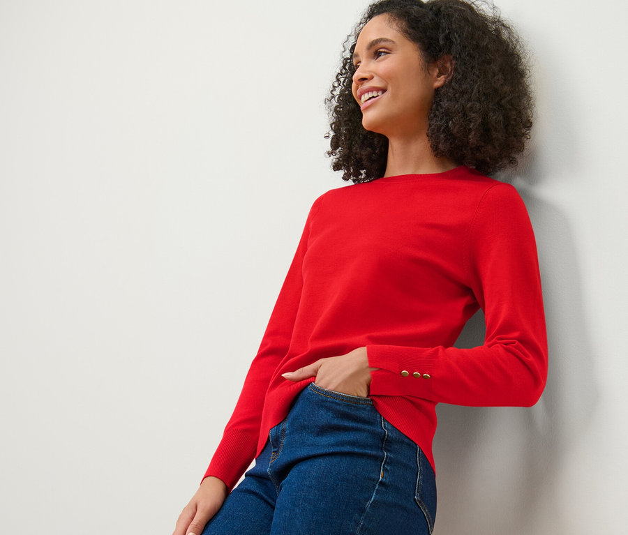 Lächelnde Frau, die sich an eine Wand lehnt, trägt einen roten Feinstrickpullover und Jeans.