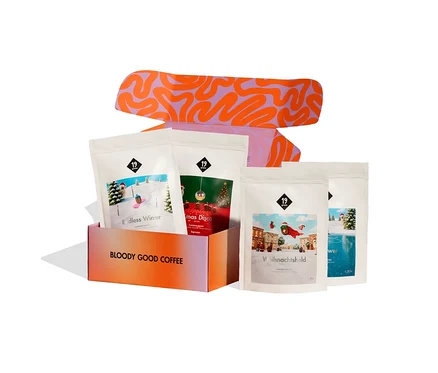 Offener 19grams Adventskalender Espresso Classic mit vier 250g Beuteln ganzen Espressobohnen. Die orange-pinke Box mit der Aufschrift „BLOODY GOOD COFFEE“ enthält zwei Beutel, zwei weitere stehen daneben. Der Deckel zeigt ein lila-orangefarbenes Wellenmuster.