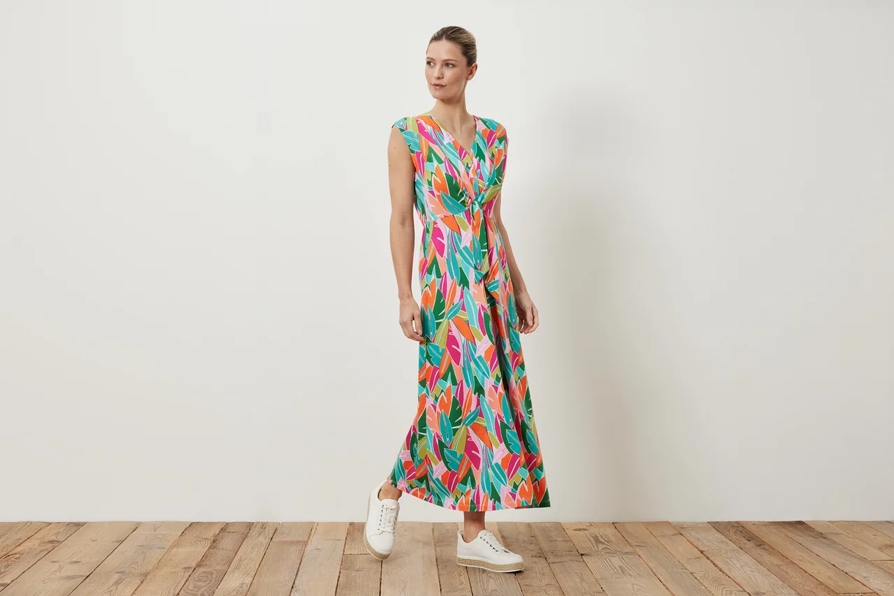 Eine Frau posiert in einem Jerseykleid mit Knoten und buntem Blattdruck, dazu trägt sie weiße Turnschuhe.