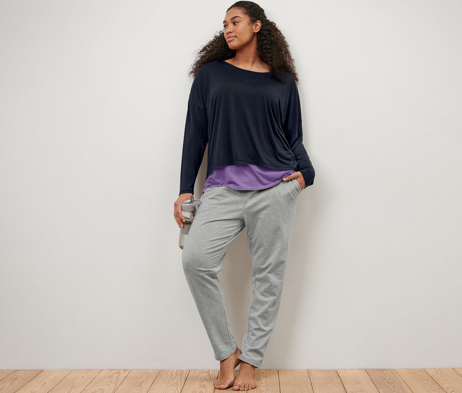 Eine Frau lehnt an einer Wand und trägt eine graue Sweathose und ein blau-lila 2-in-1-Sportshirt.