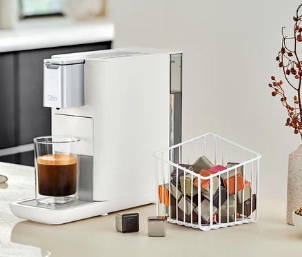 Eine Qbo ESSENTIAL Bright Sand Kaffeemaschine steht neben einem Glas Kaffee auf einer Küchentheke.