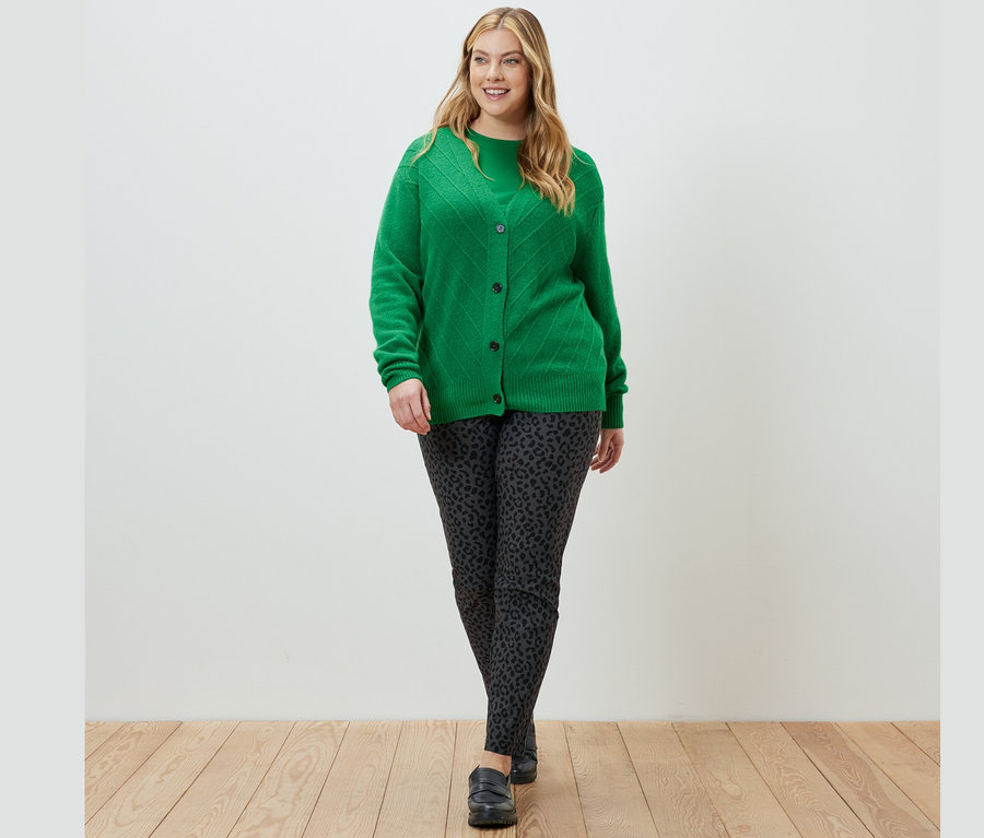 Eine Frau geht und trägt einen grünen Cardigan mit Strickstruktur, ein grünes Langarmshirt und eine bedruckte Bengalin-Hose.