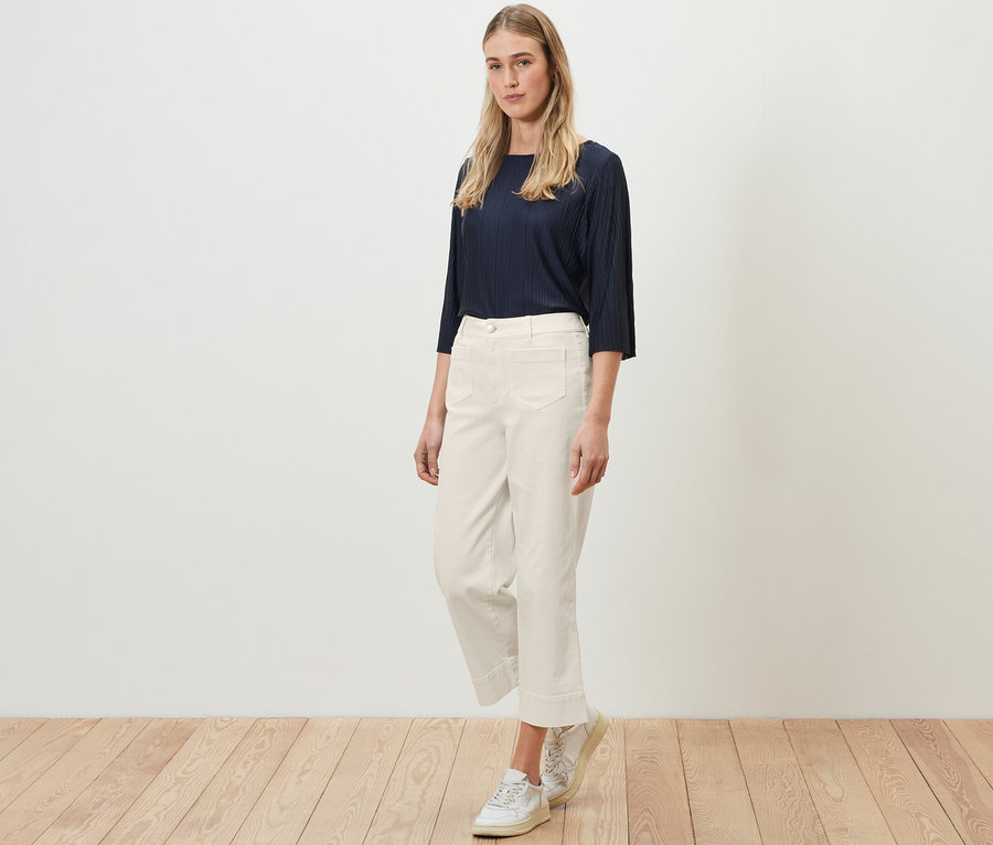Eine Frau steht auf einem Holzboden und trägt ein marineblaues Oberteil und eine cremefarbene Flared Jeans – Fit »Cara«.