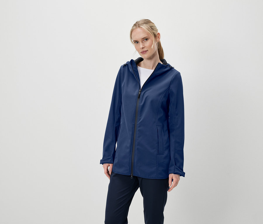 Frau posiert in einer dunkelblauen Softshelljacke mit Kapuze und dunkelblauen Hosen.