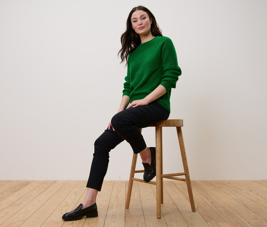 Eine Frau sitzt auf einem Hocker und trägt einen grünen Strickpullover.