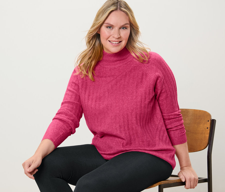 Frau sitzt auf einem Stuhl und trägt einen gerippten Strickpullover mit Stehkragen und Thermoleggings.