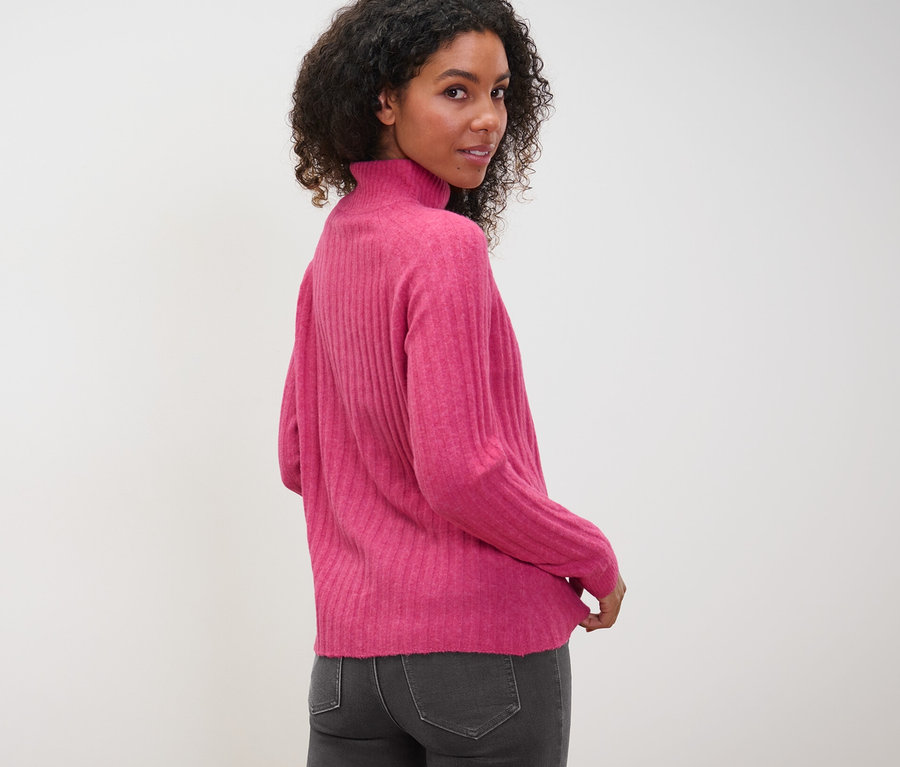 Frau blickt über die Schulter und trägt einen gerippten Strickpullover mit Stehkragen und graue Jeans.