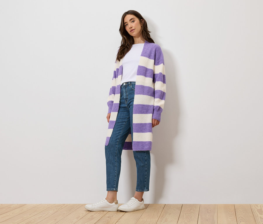 Eine Frau steht und trägt ein weißes T-Shirt, Jeans und einen lila-weiß gestreiften Cardigan.